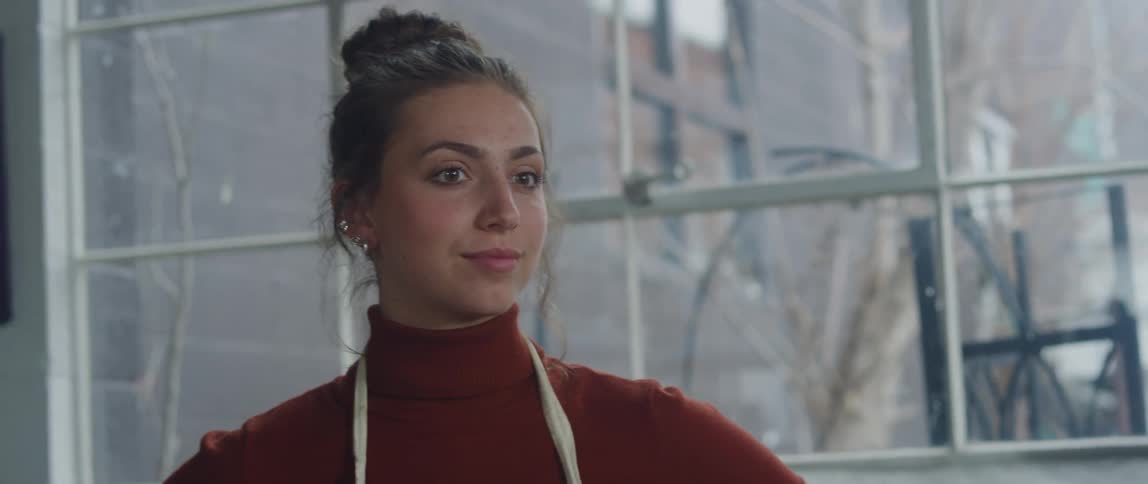 A woman with dark hair tied in a bun is wearing a red turtleneck sweater and a light colored apron as she looks at a painting with a relaxed expression