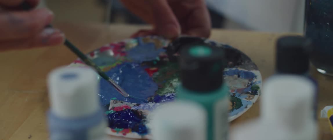 An artist mixing blue paint with a brush on a flat plate at a table with other paints in different containers placed beside it