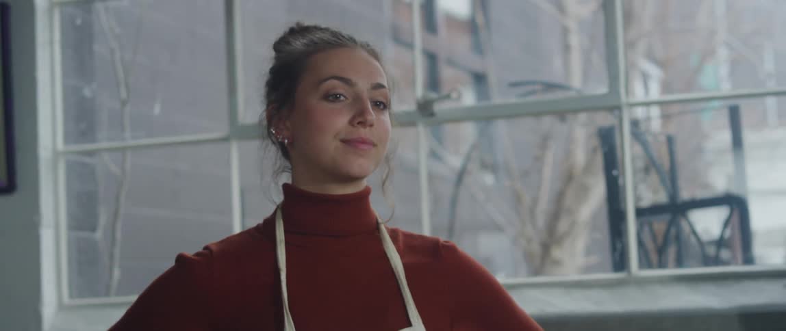 A woman with dark hair tied in a bun is wearing a red turtleneck sweater and a light colored apron as she stands next to a large window with a relaxed expression
