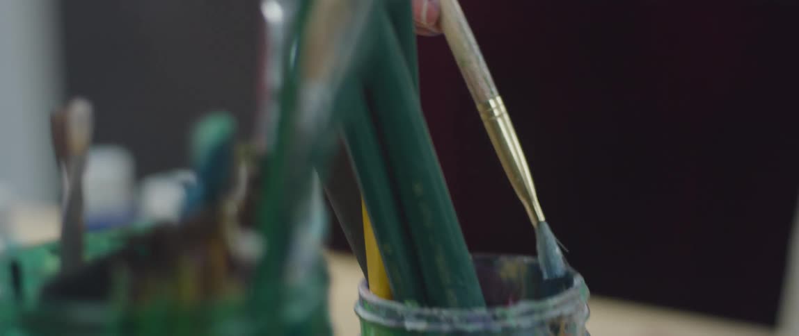A close view of an artists workbench shows a persons hand holding a paintbrush dipping it into a glass jar filled with paint brushes before pulling it out and moving it out of the frame