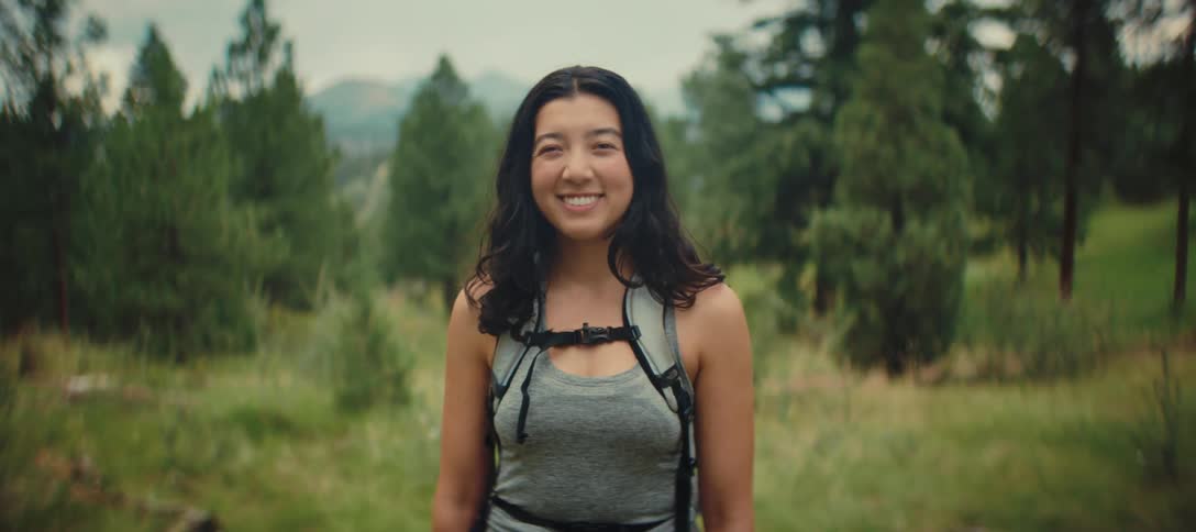 A smiling woman with a backpack stands in a sunlit forest