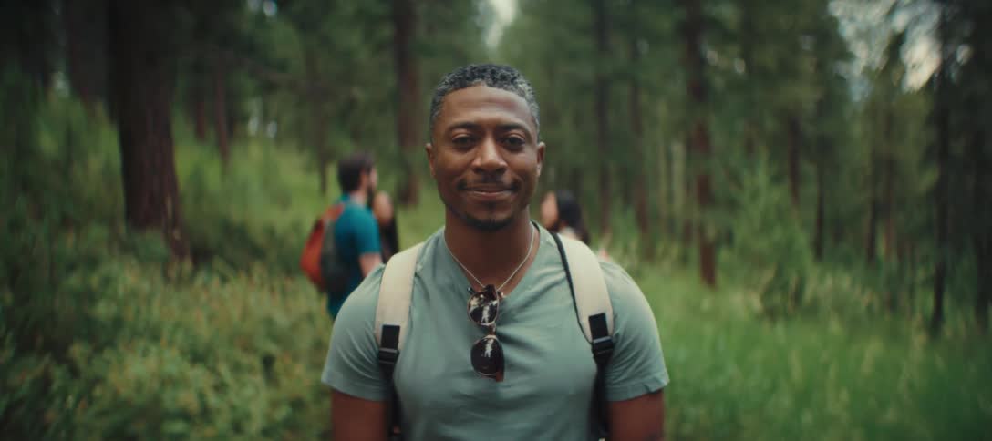 A man with a short hairstyle wearing a gray shirt and a white backpack is standing in a green forest looking directly at the camera with people visible in the background