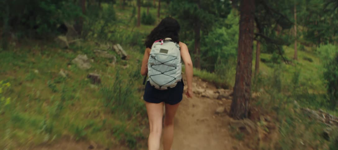 A woman wearing a light gray backpack dark shorts and hiking boots is running up a winding dirt path in a wooded area
