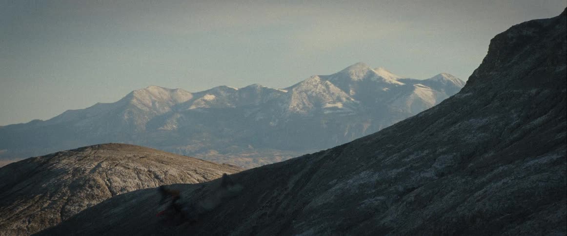 A person on a dirt bike performs a jump against a mountainous backdrop and then lands behind a hill