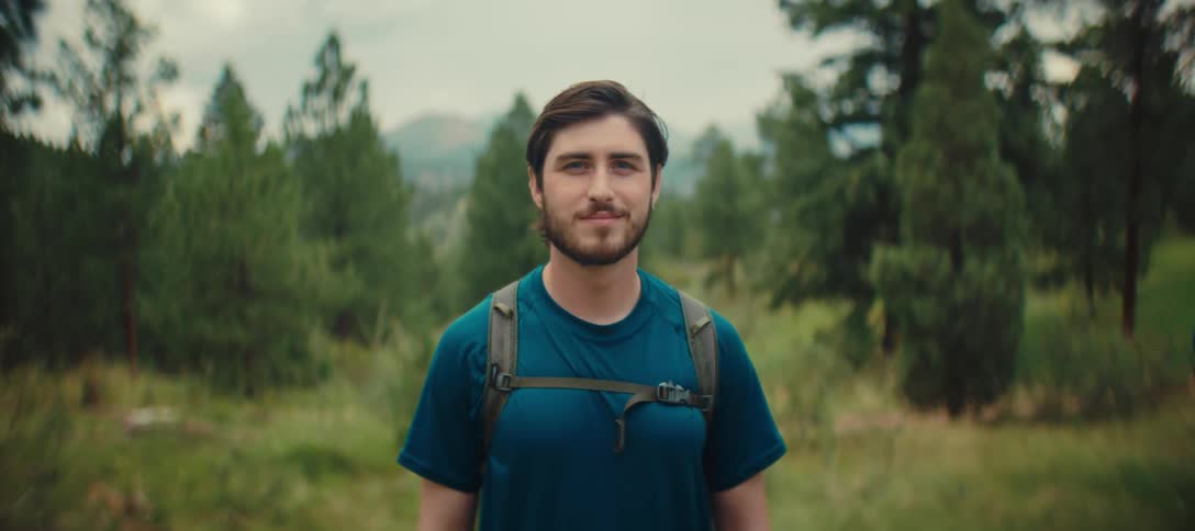 Man in a blue shirt and backpack looks forward on a trail surrounded by trees