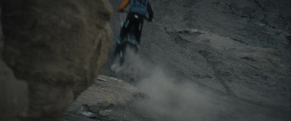 In the provided video a person on a motorbike rides down a steep rocky and barren slope as they descend a cloud of dust billows up behind them briefly obscuring the view the landscape appears rugged and desolate