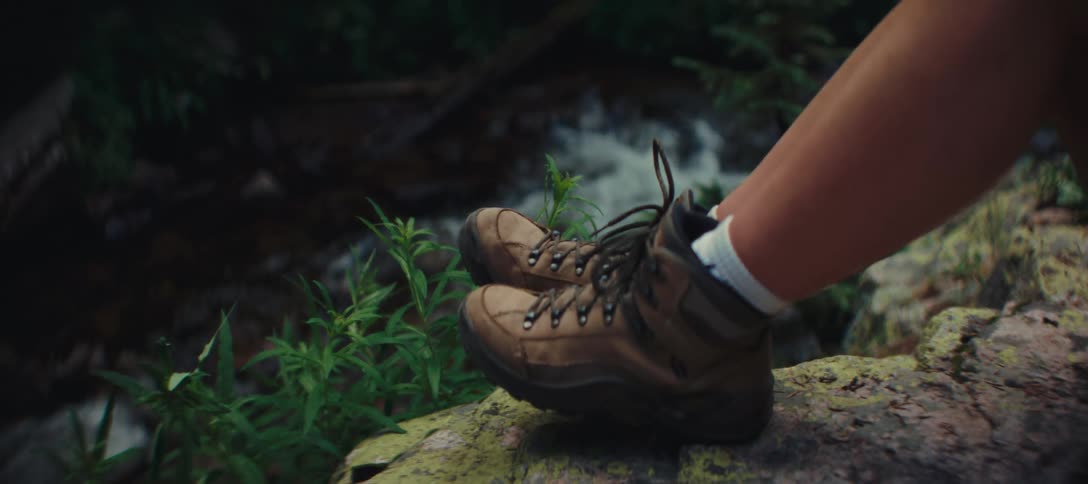 A person wearing boots sitting on a rock beside a stream in the woods at midday