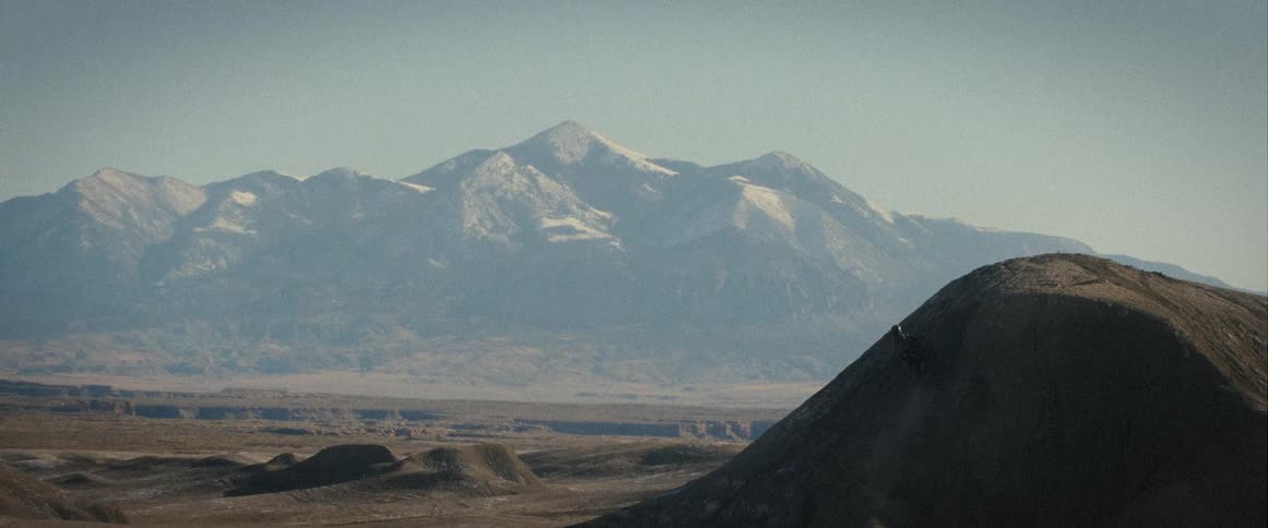 A motorbike rider jumps off a dirt hill with snowy mountains in background