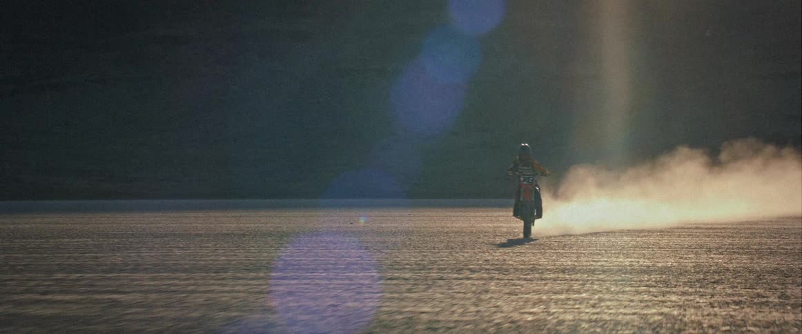 A person rides a motorcycle across a dusty flat terrain at sunset