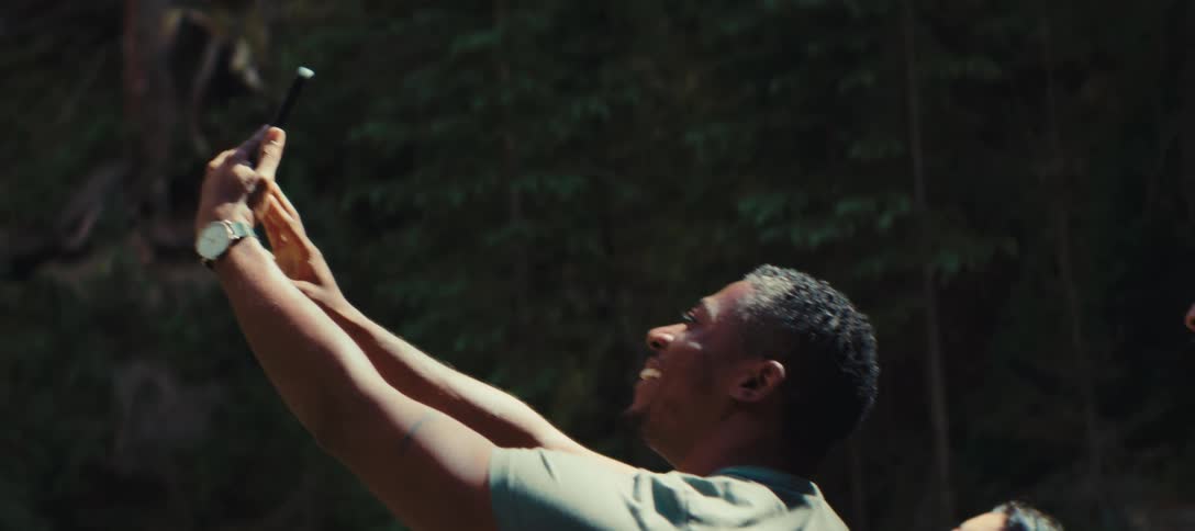 Man takes a selfie of himself and his fellow hikers while standing near a forested slope on a sunny day