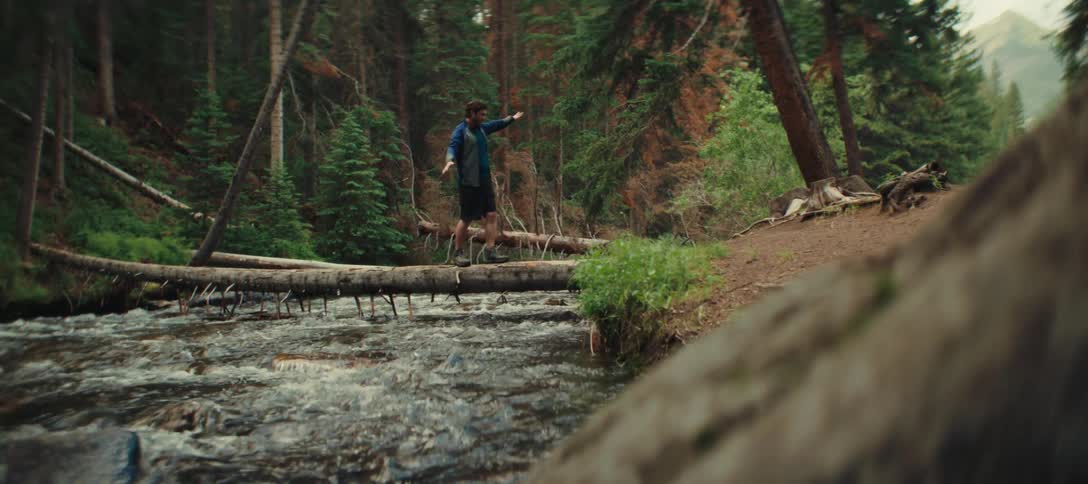 A man in a forest crossing a river by walking on a tree trunk