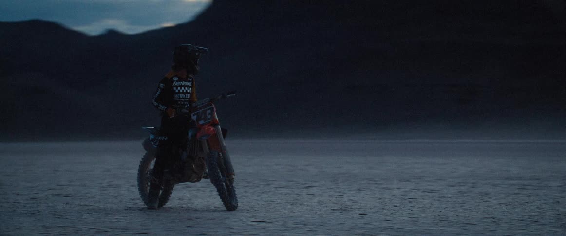 The video shows a person wearing a helmet and full protective gear standing next to a dirt bike with a mountain in the background the sky is a dark overcast blue and the scene appears to be taking place either at dusk or dawn
