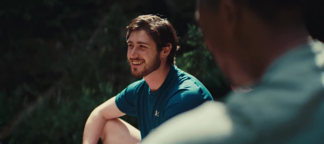 A young man sits outdoors with friends while smiling and talking