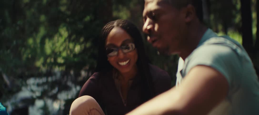 A black man and woman sit in a forest near a stream and talk happily as if being interviewed and the woman also laughs and motions with her hands