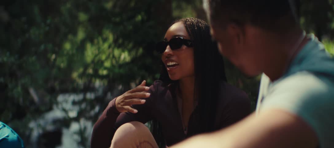 Based on the video a woman with sunglasses on is sitting outside talking and gesturing with her hands she is seated across from another person who is only partially visible
