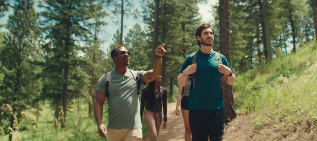 Two men walk along a forest trail with backpacks chatting animatedly