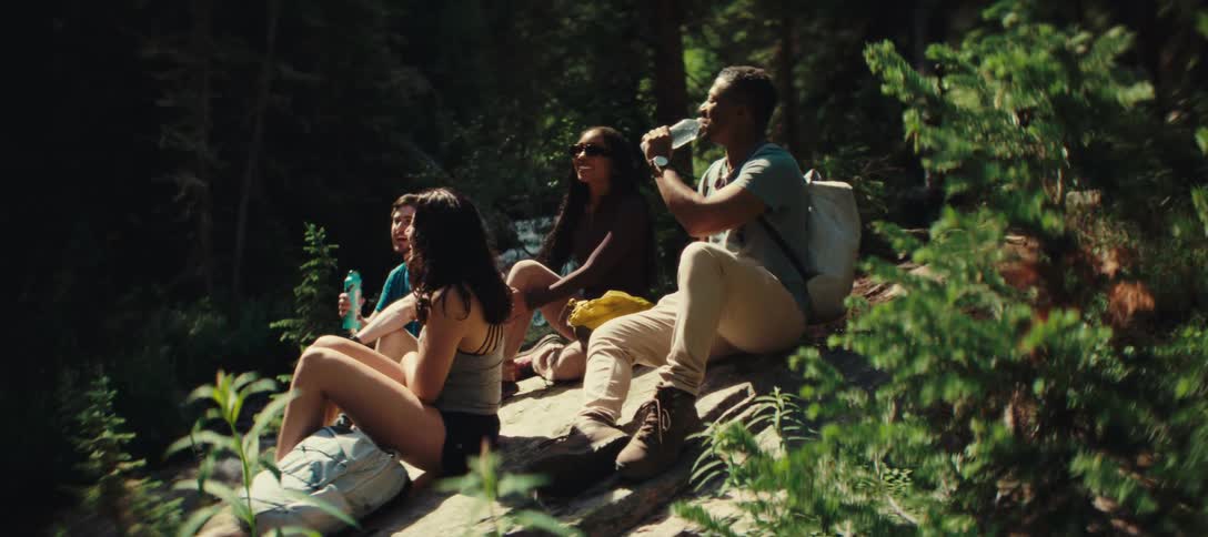 The video shows a group of four friends resting on some rocks during a hike they are sitting in a sundappled forest relaxing and sharing water from bottles