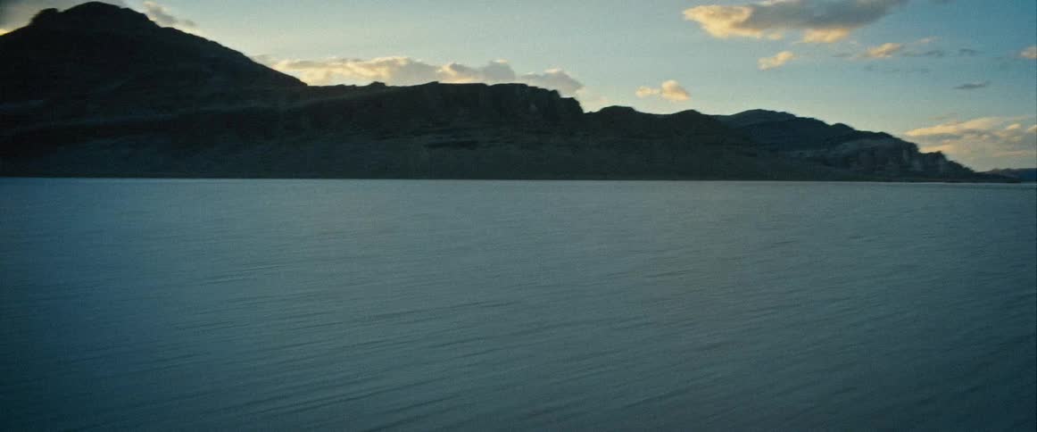A vehicle speeding across a wide flat light colored salt flat looks toward the setting sun and a large mountain range in the distance before turning backward to show a person wearing a helmet and a blue and yellow racing jersey riding a motorcycle
