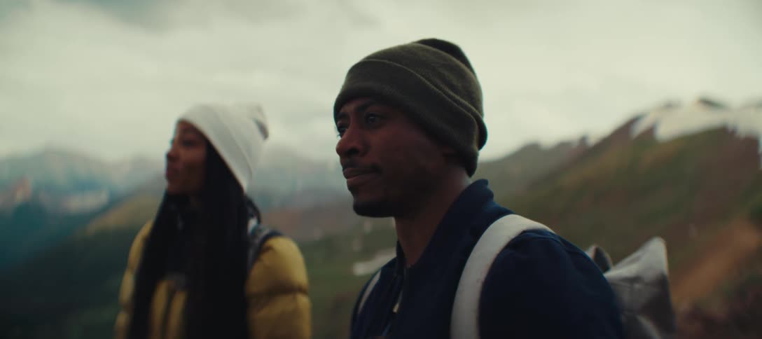 Man wearing a beanie and backpack looks at the mountain landscape beside a companion