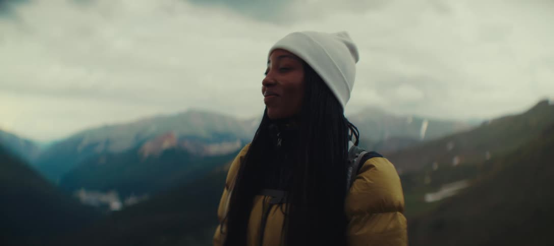 A woman wearing a coat and a winter hat at the top of a mountain taking a deep breath and smiling slightly