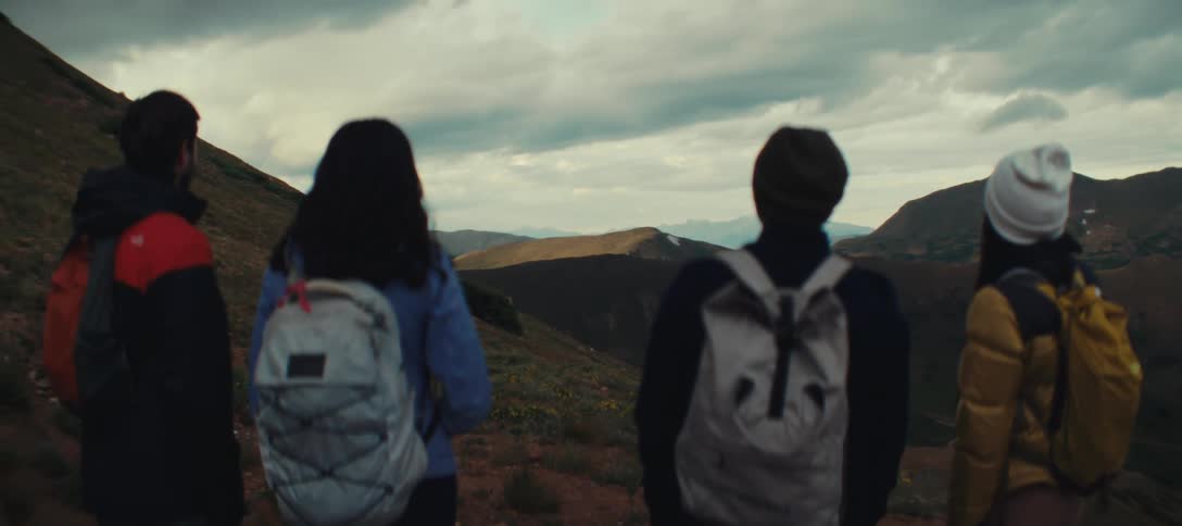Four people with backpacks stand with their backs to the camera looking out over a mountainous landscape under a cloudy sky