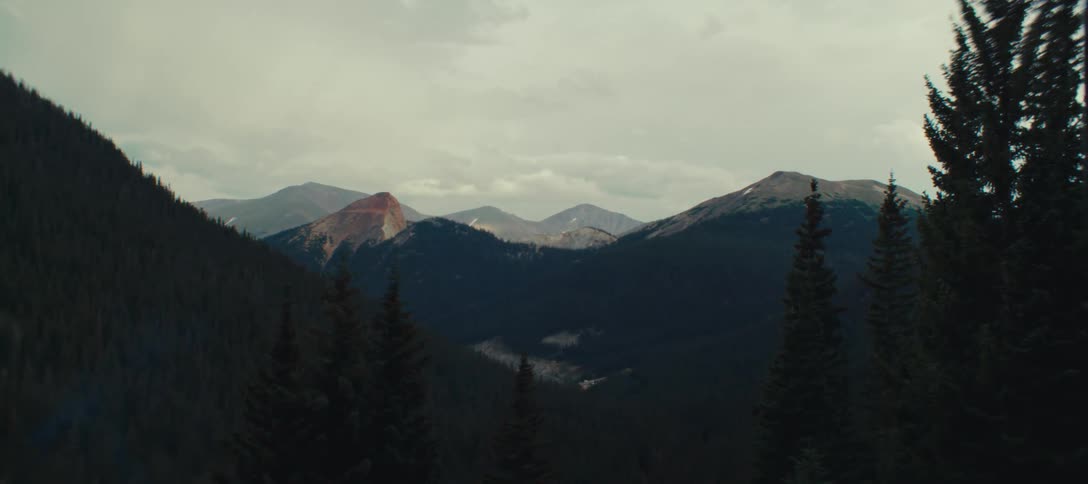 A camera pans down from a view of distant mountain peaks to reveal a deep forested valley below under a cloudy sky