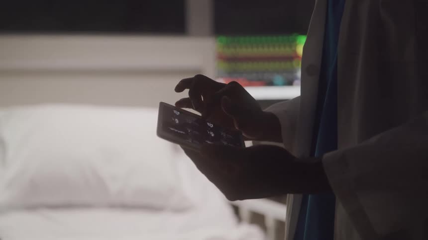 A doctor or member of the medical staff is standing in a room and medical equipment in the background using their fingers to scroll through a tablet held in both hands