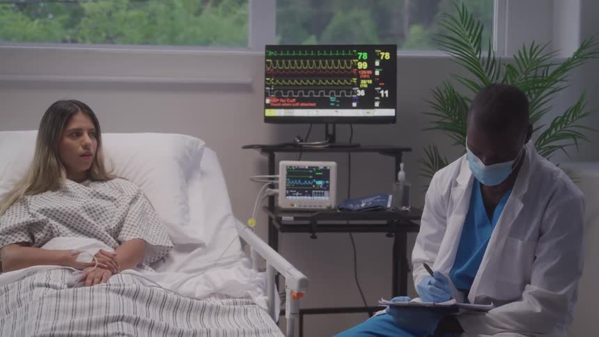 A woman lying in a bed talking to a doctor who is sitting nearby taking notes
