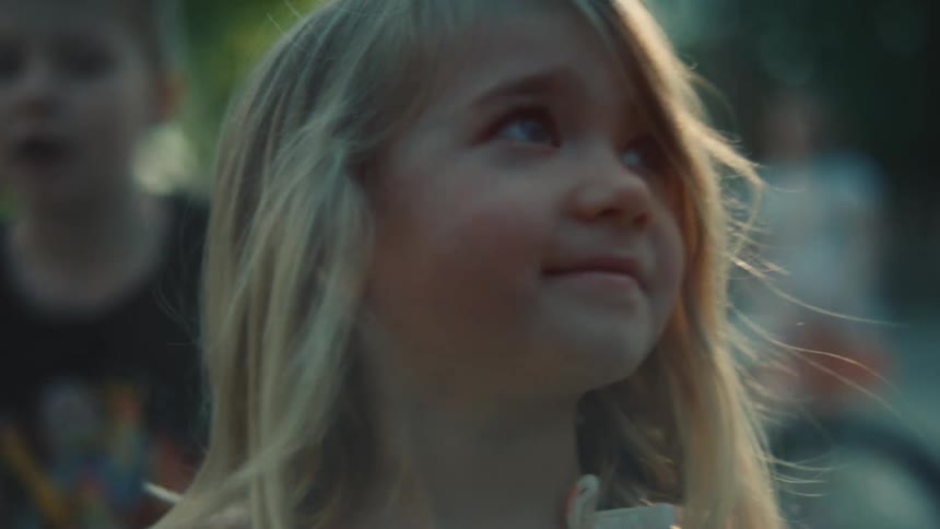 The video shows a closeup of a young girl with blonde hair who is slightly smiling the camera moves in a circular motion around her as she looks off to the side there are other children and what appears to be a backyard in the background
