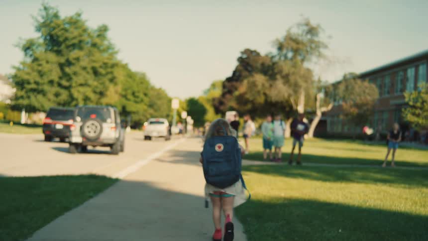 Young girl wearing a backpack walks along the sidewalk while approaching school on a sunny day