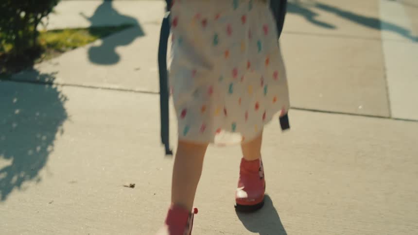 A child walks on the sidewalk at midday carrying a backpack with other kids behind her who seem to be coming from school