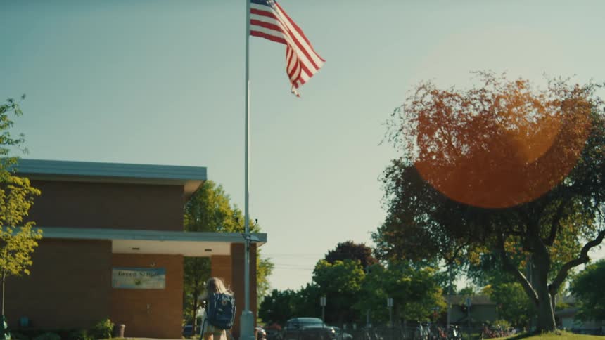 A child running toward a school building on a bright sunny day with the main focus being a large american flag waving prominently in the wind on a flagpole in the foreground