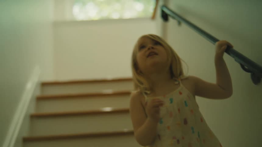 A little girl with light colored hair and a white dress with a colorful pattern is walking down a staircase holding onto a dark wooden handrail