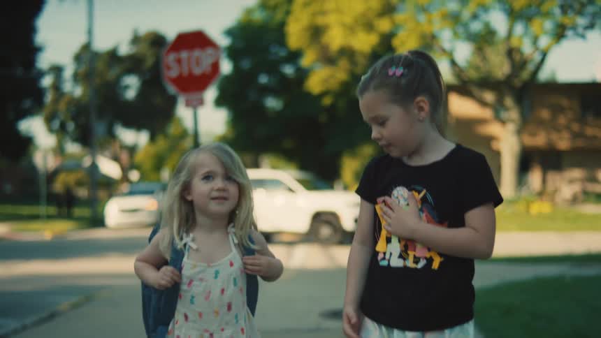 Two young girls walk together on a sidewalk in a suburban neighborhood