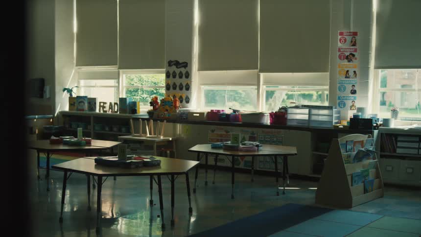 A view of an empty classroom shows a variety of play kitchen toys and furniture arranged on a gray floor with several large windows and white blinds lining the far wall