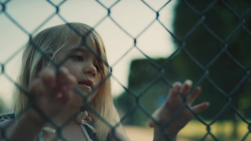 A child stands in a field at midday playfully holding a wire mesh fence with both hands