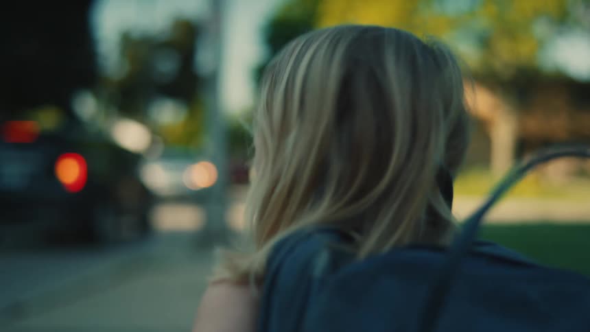 The video shows a young girl with blonde hair seen from behind as she walks down a street she is wearing a dark blue backpack and turns to look over her shoulder