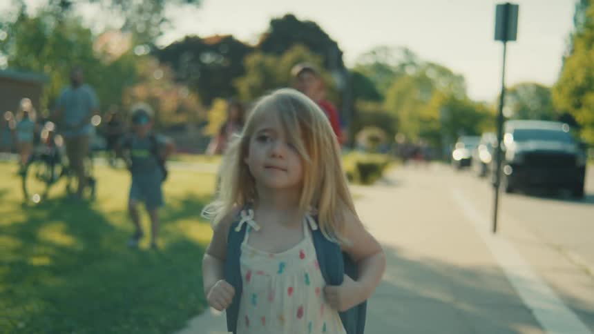 A small girl with a backpack is walking on a sidewalk she has long blonde hair and is wearing a colorful dress in the background other people and kids are visible some on bicycles