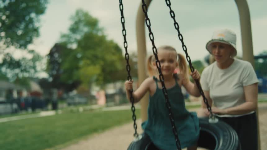 A young blonde girl in pigtails is pushed on a tire swing by a woman in a bucket hat at a park