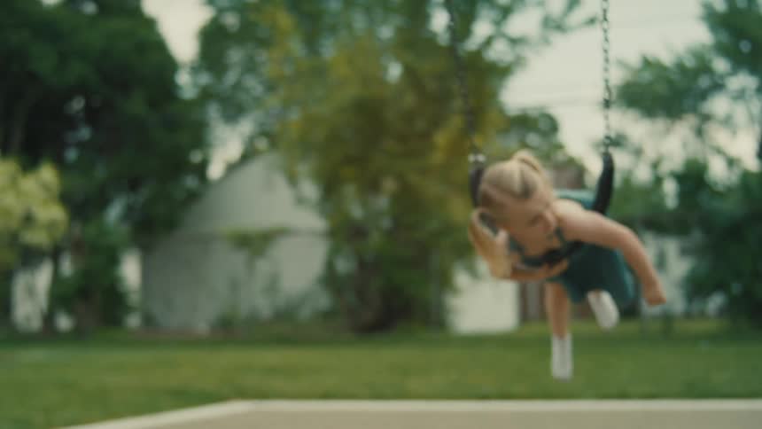 A little girl hangs face down on a swing in the playground at midday swinging back and forth with a smile on her face