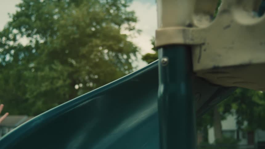 A girl wearing red shoes and a blue dress plays on a plastic playground walking across a narrow angled bridge before descending a curving blue slide