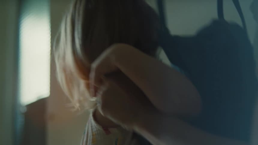 A person helps a young blonde girl put on a dark backpack then the girl turns and walks towards a sunlit doorway