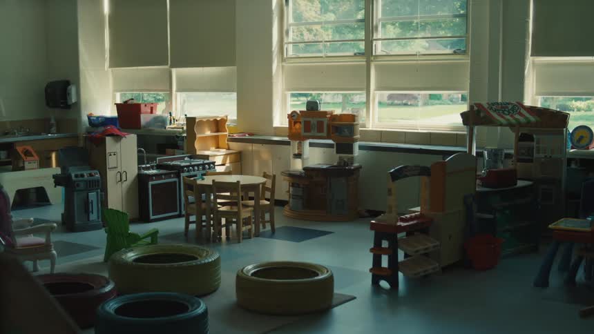 A view of an empty classroom shows a variety of play kitchen toys and furniture arranged on a gray floor with several large windows and white blinds lining the far wall