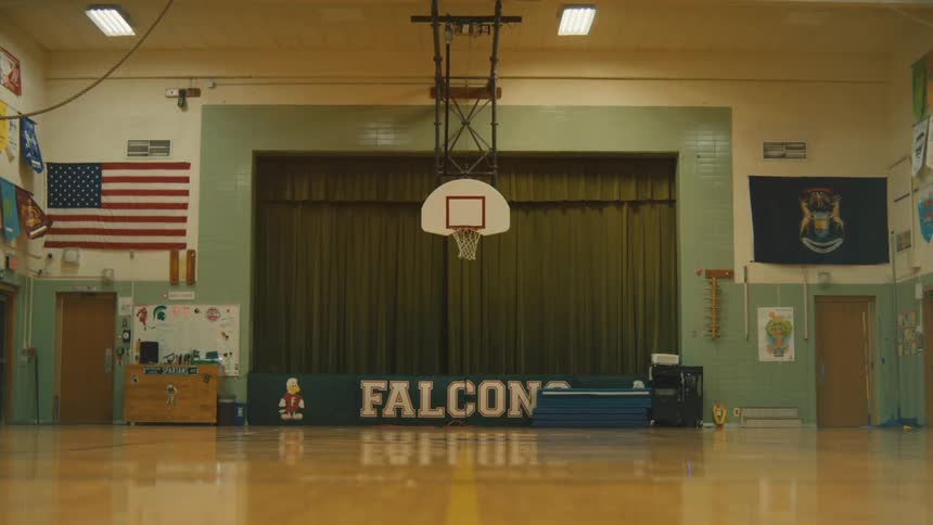 The video shows an empty school gymnasium in the center of the frame there is a stage with a dark green curtain and a banner that reads falcons a basketball hoop hangs from the ceiling in front of the stage