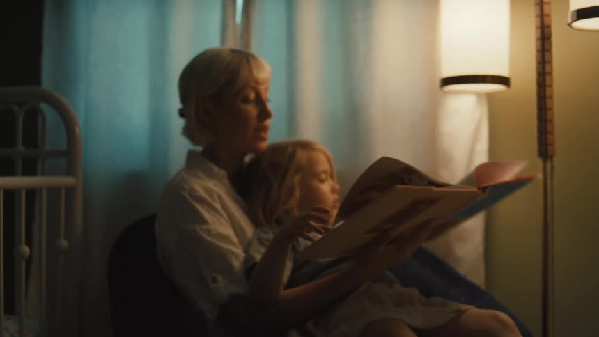 A mother and daughter are sitting together in a chair the mother is reading a book to the daughter who is pointing at something on the pages they are sitting in a dimly lit room with a tall lamp providing light