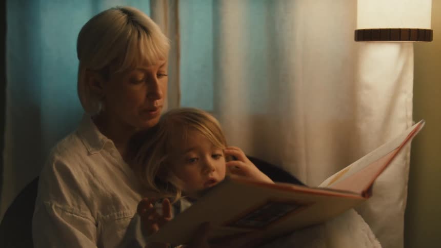 A blonde woman holds a young blonde child in her lap reading a book to them under the warm glow of a lamp