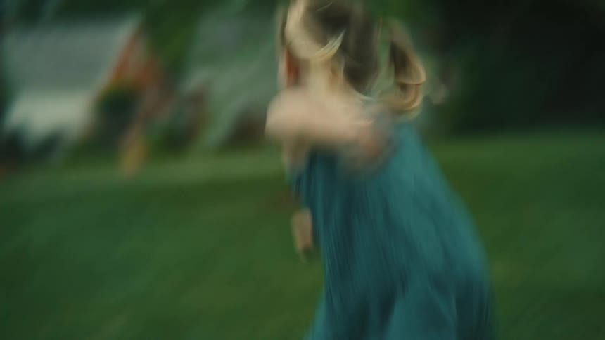 A child with pigtails running joyfully across a green lawn as a woman with a bucket hat playfully bends down and encourages them
