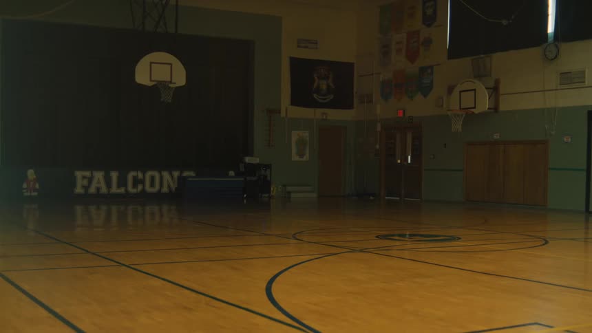 An empty school gymnasium with a hardwood court basketball hoops and banners on the walls is shown with dim lighting
