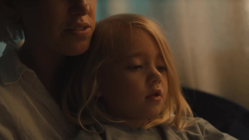 The video shows a closeup of a mother and her young child both with blonde hair sitting together in a dimly lit room the mother is holding a book and reading aloud while the child looks on intently captivated by the story