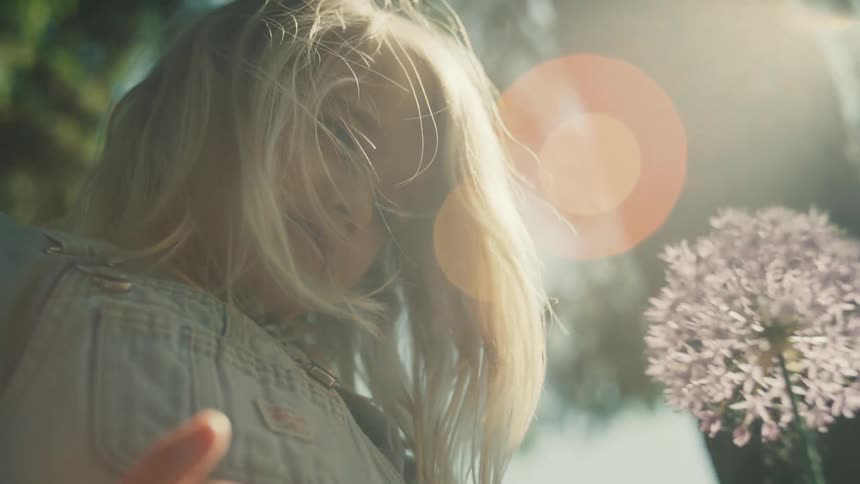 A young blonde haired girl in overalls gently touches a purple flower while bright sunlight creates a lens flare